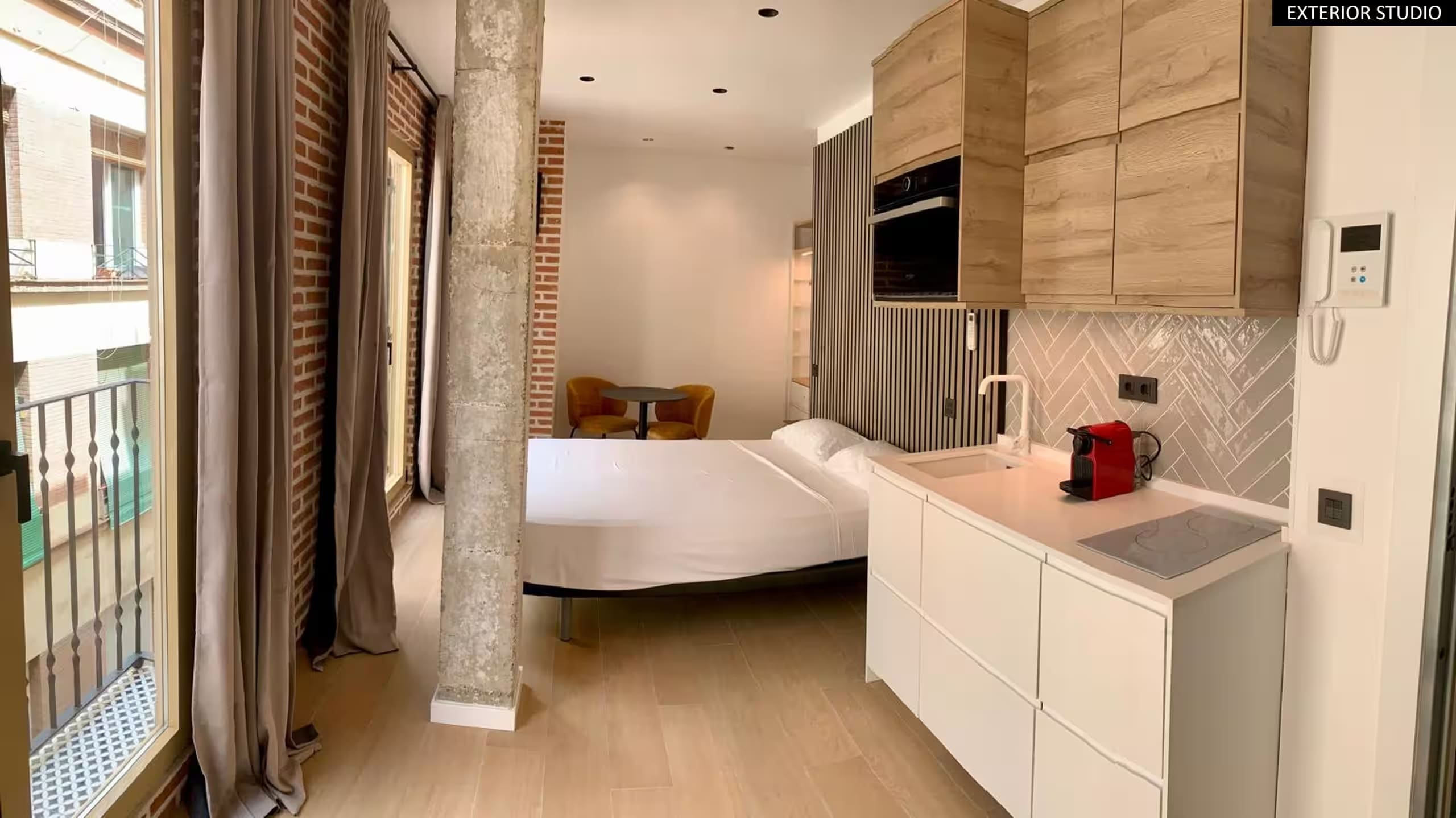 Estudio en Málaga antes del retoque: cama, cocina integrada y pared de ladrillo visto junto al balcón