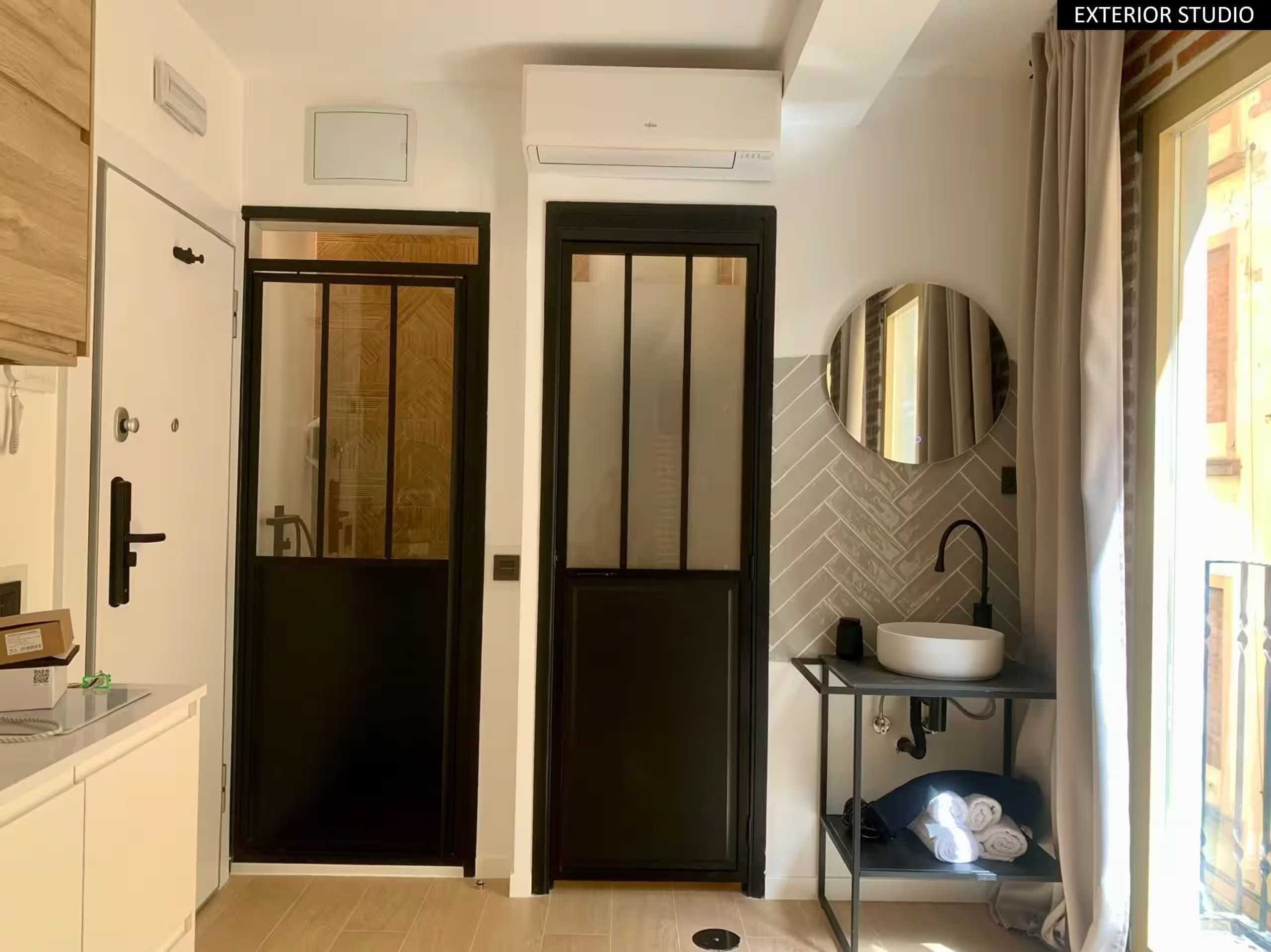Entrada y zona de baño del estudio de Málaga antes del retoque, con lavabo, espejo redondo y puertas acristaladas